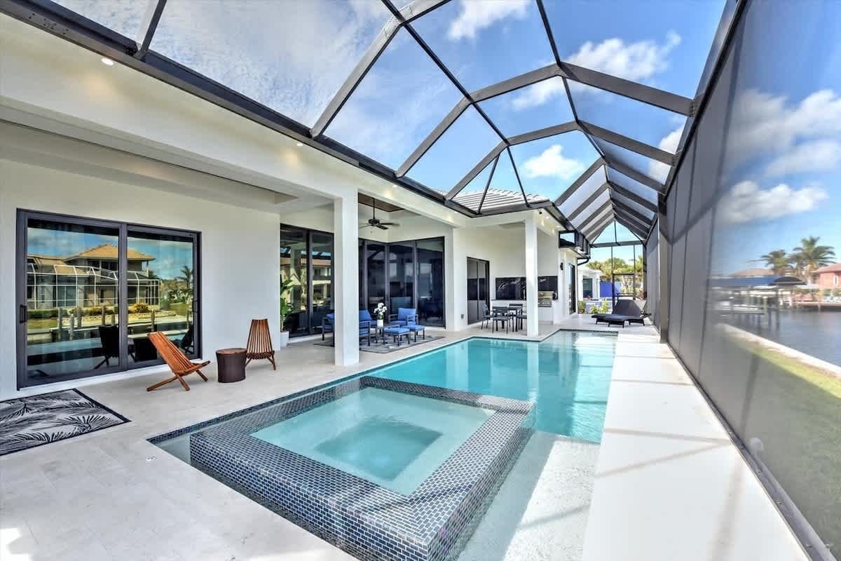 Un área de piscina interior moderna con paredes de vidrio, vistas despejadas al exterior y una zona de estar con sillones y mesas.