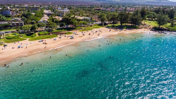 Vista aérea de una playa con agua turquesa cristalina, una orilla arenosa y edificios residenciales y colinas al fondo.