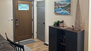 Een kamer met een houten deur, een grijze deur, een schilderij van een berglandschap, een decoratieve kegel en een kast met lades.