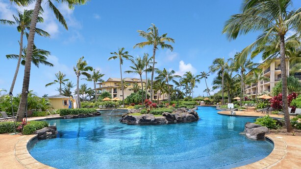 Piscina de resort con palmeras, sillones de descanso y un edificio con balcones.