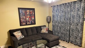 A living room with a sectional sofa, a glass coffee table, patterned curtains, a framed artwork, and a patterned rug.