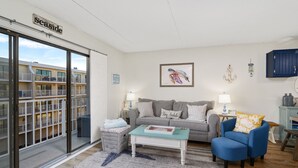 Una sala de estar con un sofá gris, un sillón azul, una mesa de centro y vistas a un edificio con balcones.
