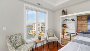 Un dormitorio con un ventanal grande, una cama, una estantería de madera y dos sillones estampados.