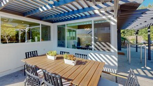 An outdoor patio with a wooden table and chairs, large windows, and a blue pergola.