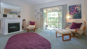Un salon avec une cheminée, deux fauteuils, un tapis rond de couleur violette et une grande fenêtre avec des rideaux.