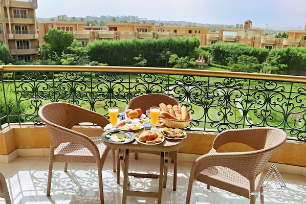 A balcony with wicker chairs, a table with breakfast items, and a view of buildings and greenery. - Grand Museum Pyramids Inn (Kirdasah)