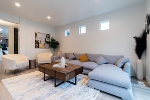 A modern living room with a grey sofa, white armchairs, a wooden coffee table, and a decorative mirror on the wall.