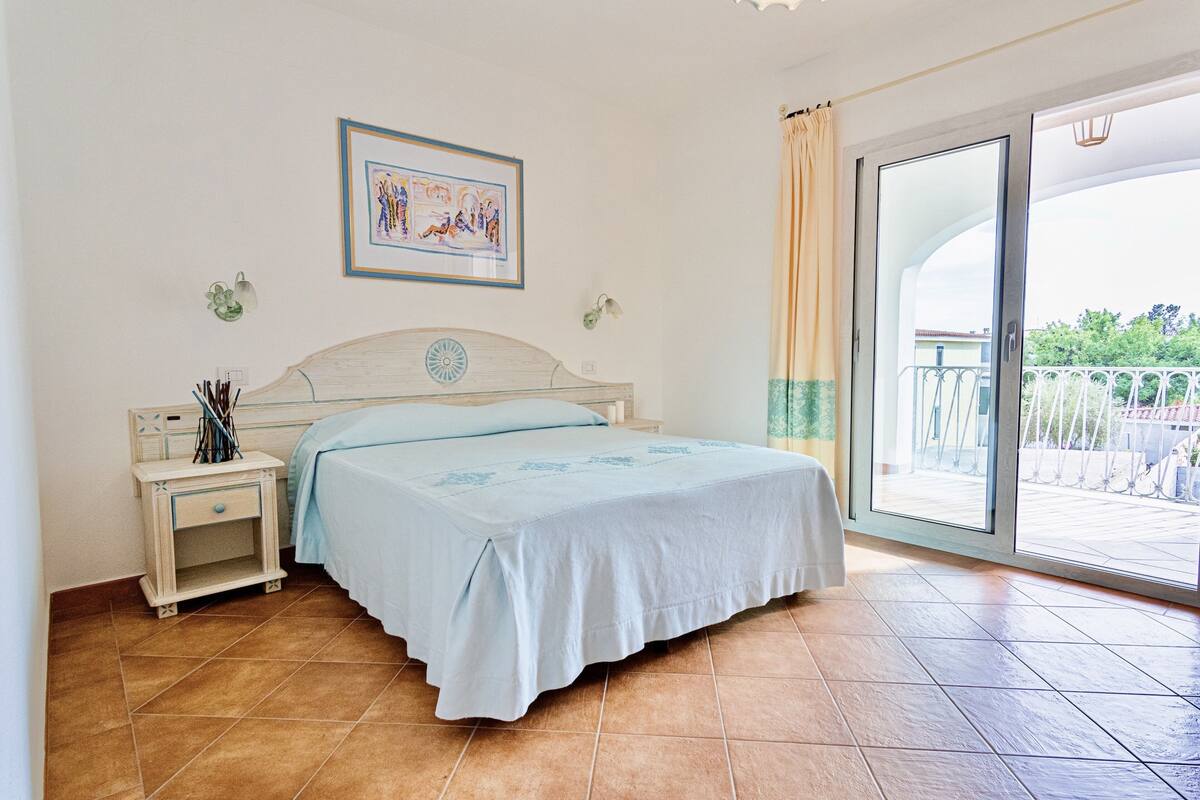 A bedroom with a bed, a nightstand, a framed picture, and a sliding glass door to a balcony.