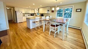 A modern kitchen with white cabinets, a central island, and a dining area with four chairs. - Coveside: Town Cove Waterfront (Orleans)