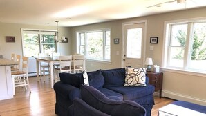 A living room with a dark blue sofa, a dining table with chairs, and large windows with natural light. - Coveside: Town Cove Waterfront (Orleans)