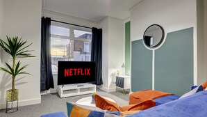 Un soggiorno moderno con una grande televisione a schermo piatto che mostra 'NETFLIX', una pianta in vaso, uno specchio rotondo e un divano con cuscini colorati.