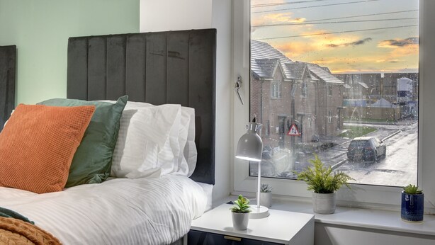 Una camera da letto con un letto, cuscini, un comodino con una lampada e una finestra vista strada e palazzi.
