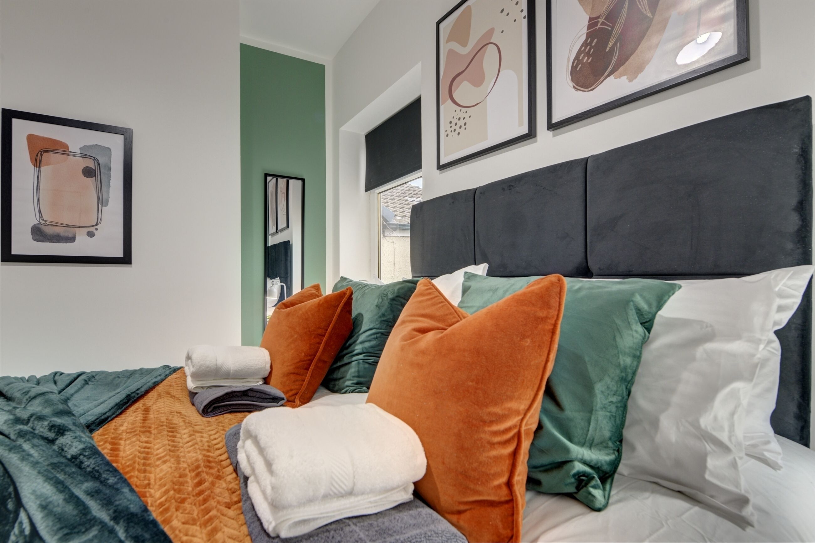 A neatly made bed with multiple pillows and a blanket, framed artwork on the wall, and a window with a view.