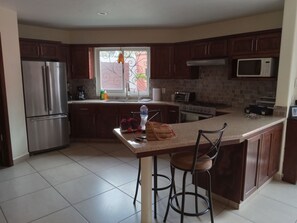 A modern kitchen with stainless steel appliances, a large island with bar stools, and a view through a window with decorative ironwork. - Beautiful New Casita-Ajijic West 360 Degree View of Lake/Mountains (Ajijic)