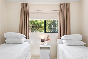 Two single beds with white bedding and matching pillows, a small nightstand with a lamp and flowers, and a window with a view of greenery.