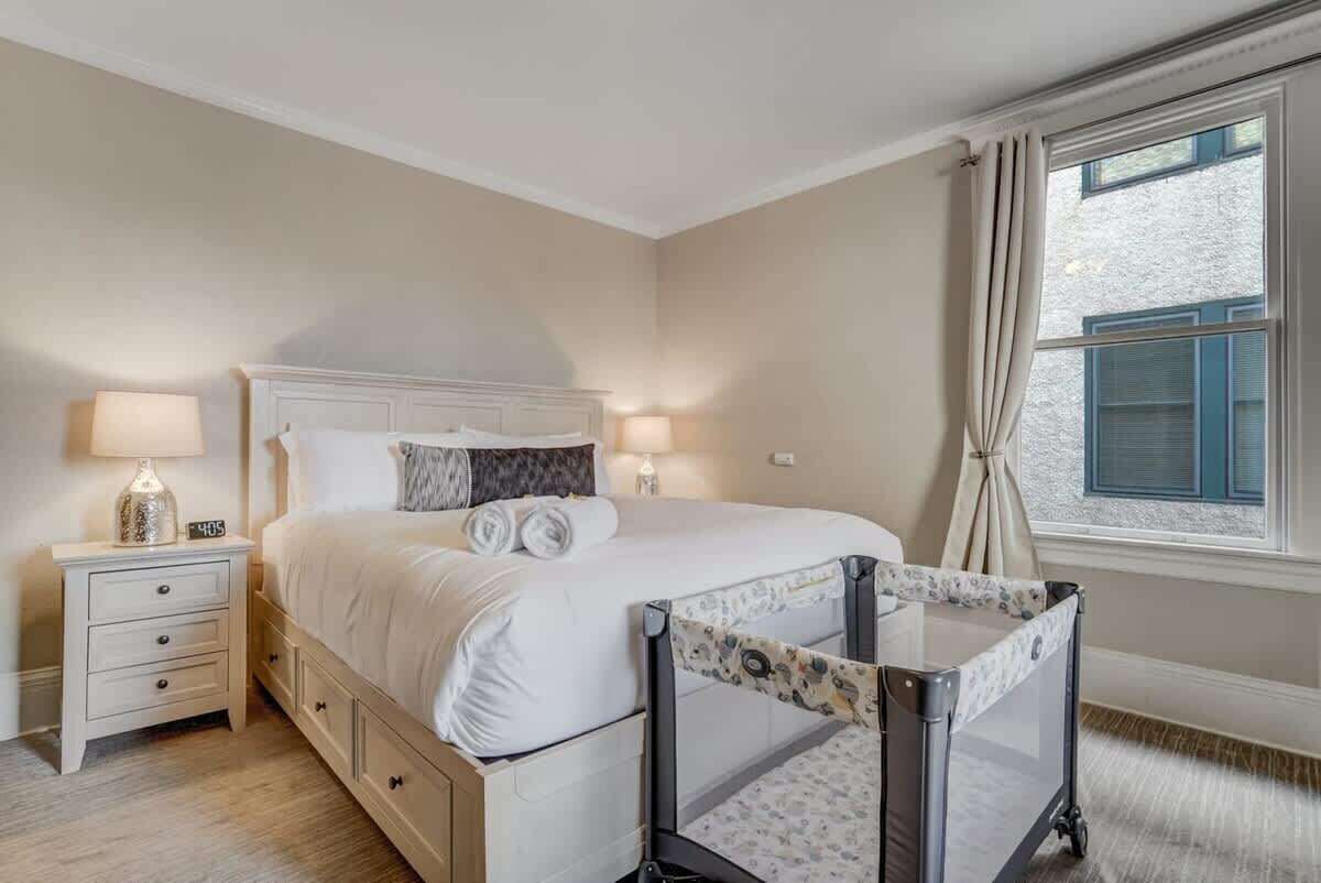 A bedroom with a bed, bedside lamps, a nightstand, a window with curtains, and a crib.