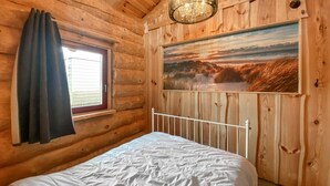 A wooden cabin bedroom with a bed, a window with curtains, and a painting on the wall. - Unique log house for 6 pers. with terrace and unobstructed view near Wadden Sea (Den Oever)