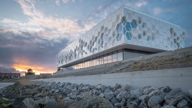 A modern building with a geometric facade, large windows, and a rocky shoreline. - Unique log house for 6 pers. with terrace and unobstructed view near Wadden Sea (Den Oever)