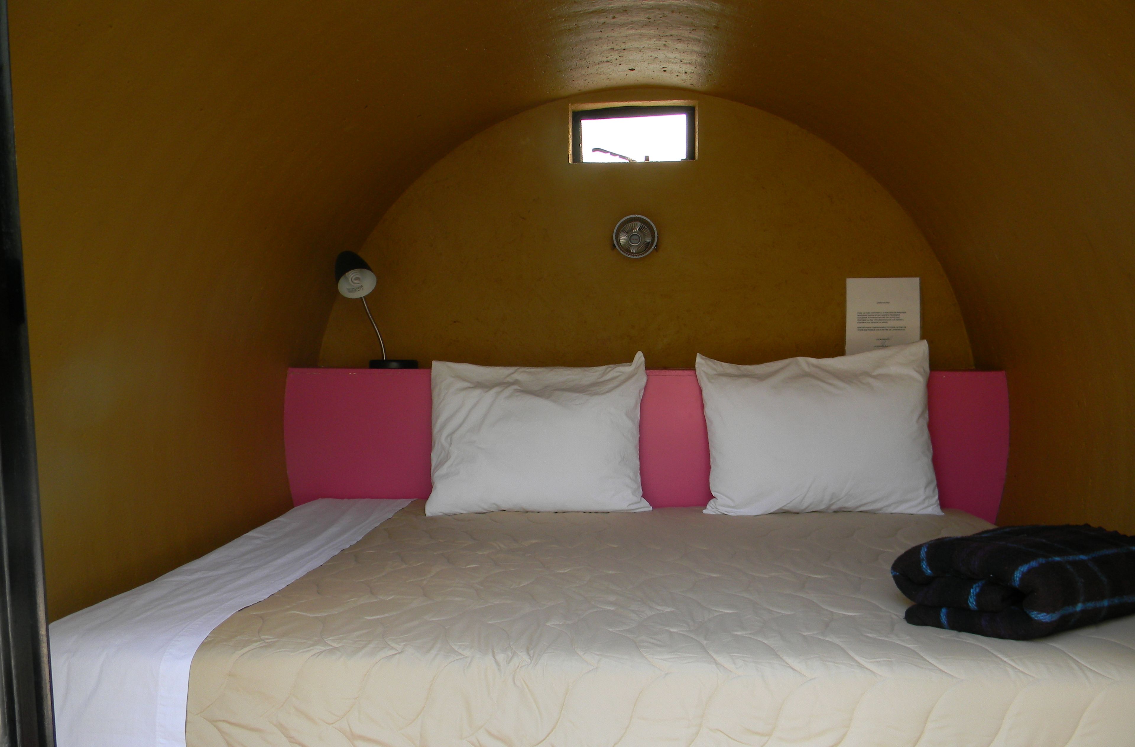 Una cama individual con cabecera rosa y ropa de cama blanca, una ventana pequeña encima de la cama y un reloj de pared.