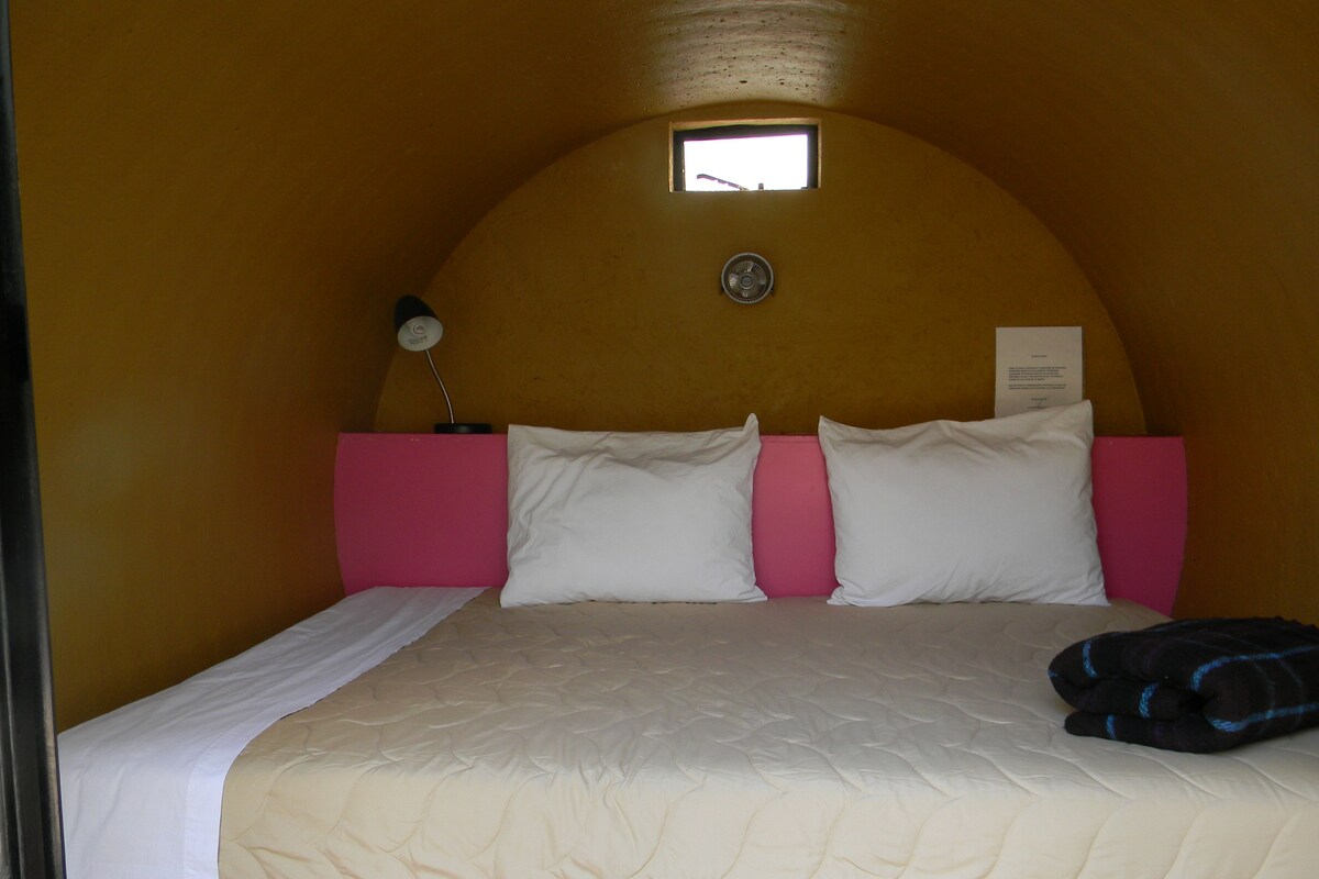 Una cama individual con cabecera rosa y ropa de cama blanca, una ventana pequeña encima de la cama y un reloj de pared.