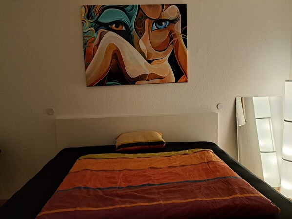 Een bed met een kleurrijk gestreept dekbed, een kussen en een schilderij aan de muur erboven.