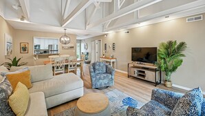 A spacious living room with a sofa, armchairs, a coffee table, and a TV. There is a kitchen in the background with cabinets and a dining area.