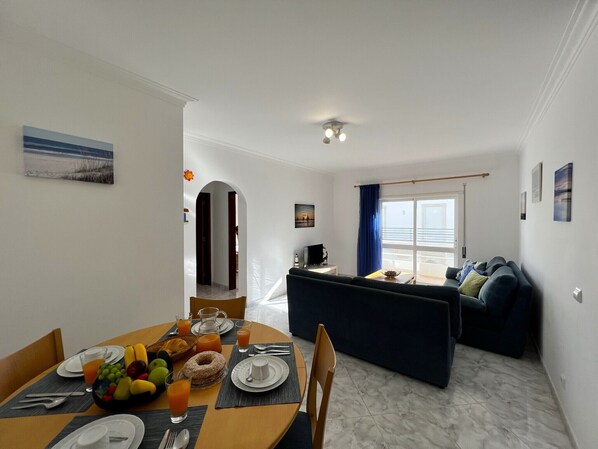 A modern living room with a dark blue sofa, a dining area with a breakfast setup including fruit and juice, and a balcony with a view.