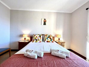 A hotel room with a large bed, two bedside tables with lamps, a small table with a clock, and a wall-mounted picture.
