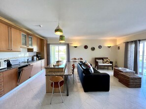 A modern kitchen with granite countertops, wooden cabinets, and a central island. There is a dining area with a black sofa and a coffee table. The room has a large window with curtains, and a ceiling fan is visible.