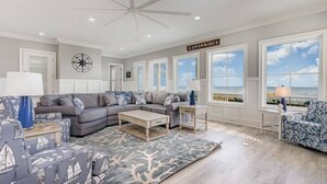 Ein großzügiges Wohnzimmer mit einer großen, grauen Eckcouch, einem Couchtisch und einem gemusterten Teppich. Zwei Nachttische mit Lampen, ein Deckenventilator und ein großes Fenster mit Meerblick.