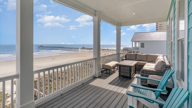 Eine überdachte Veranda mit Blick auf Strand und Steg, möbliert mit Sitzgelegenheiten und einem Tisch.