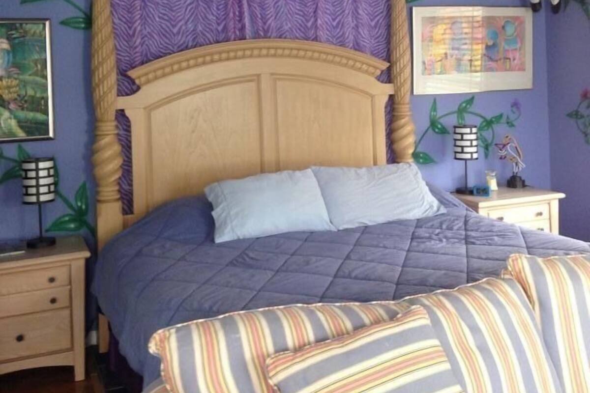 A bedroom with a wooden headboard, a bed with a purple quilt, striped pillows, and a nightstand with a lamp.