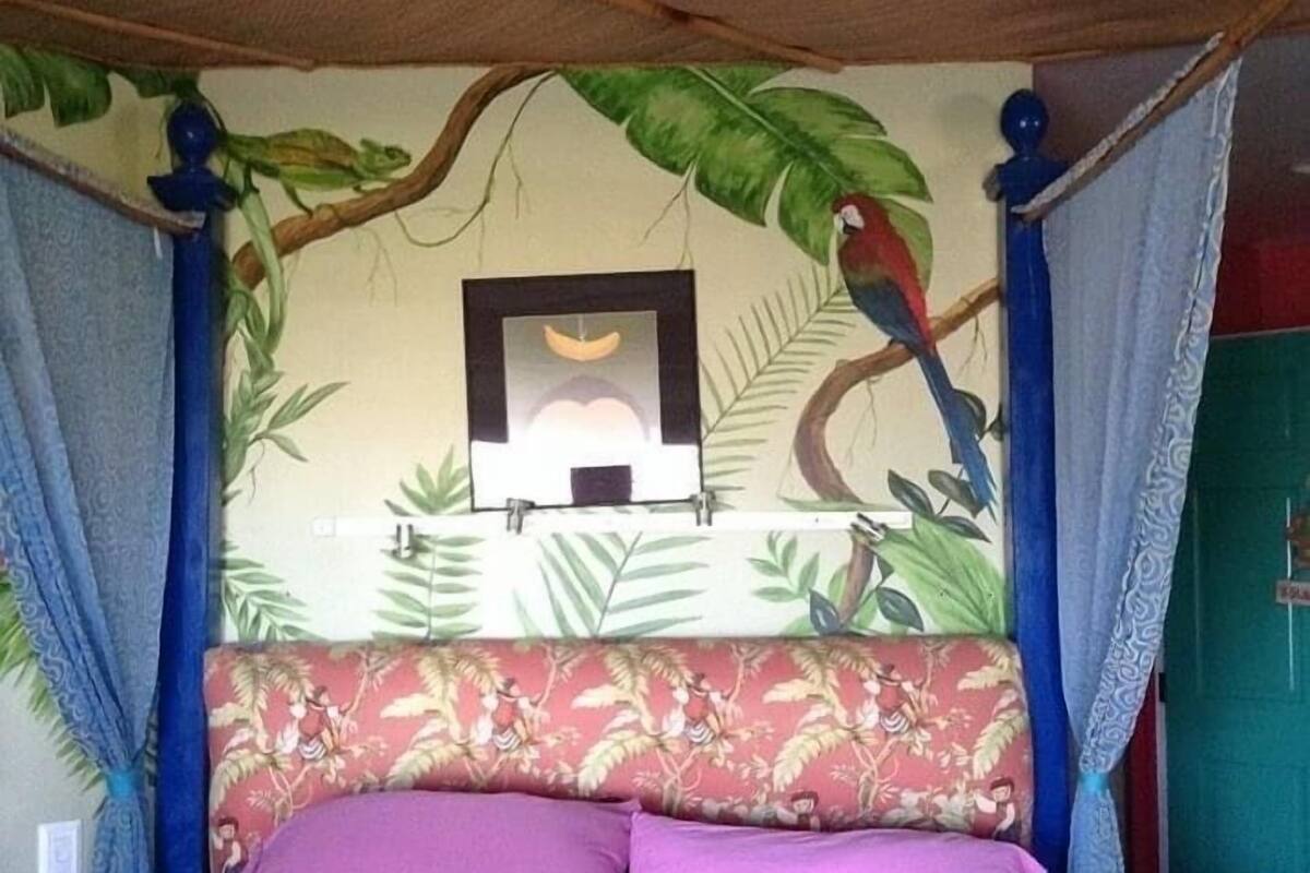 A bed with a canopy, a wall mural with a bird and foliage, and a framed picture on the wall.