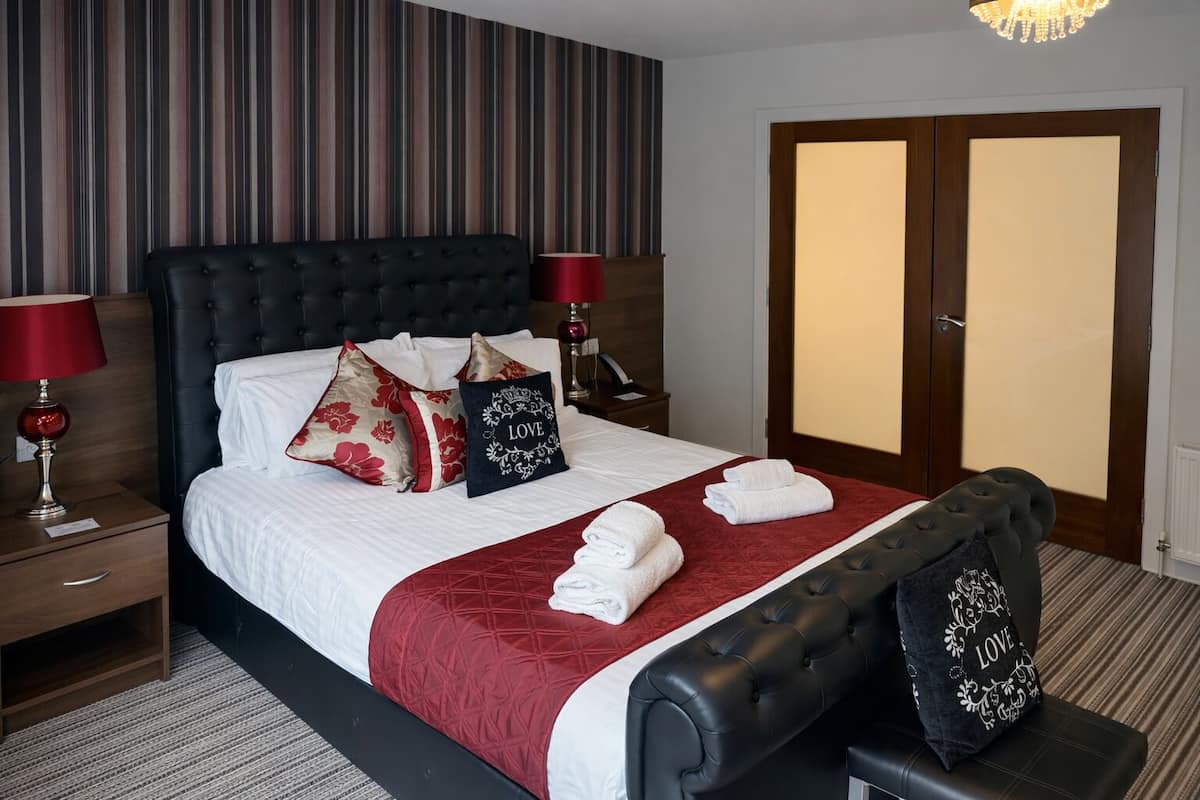 A hotel room with a large bed, red and white bedding, a dark headboard, two bedside tables with lamps, a red throw blanket, and a bench with a pillow.