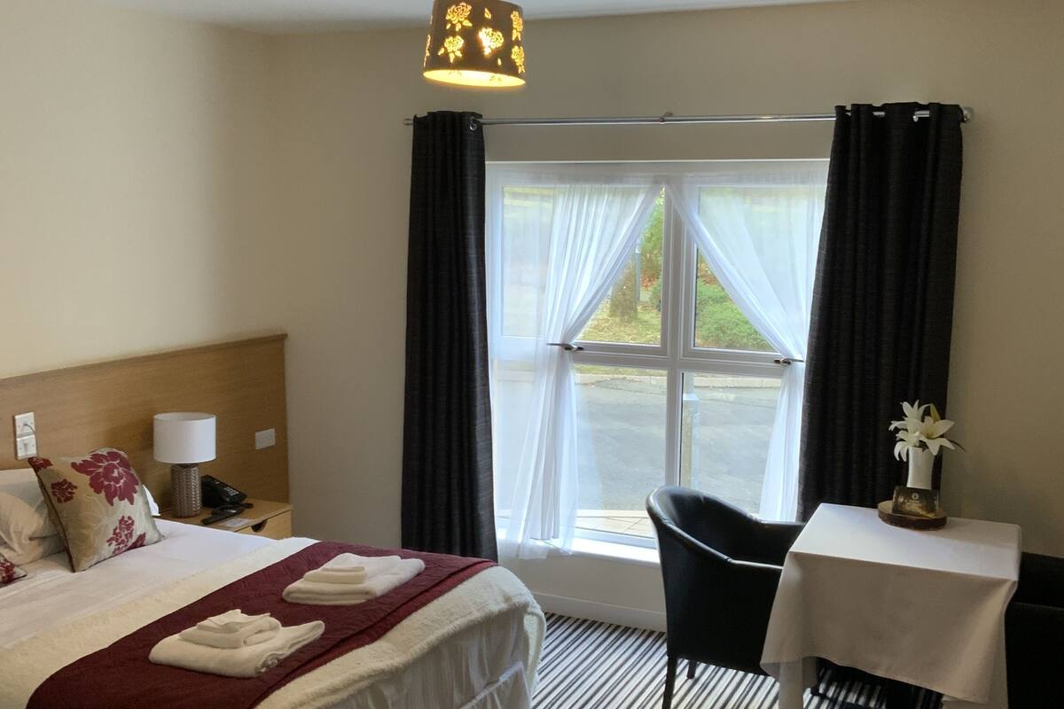 A hotel room with a bed, a desk, a chair, a lamp, and a window with curtains.