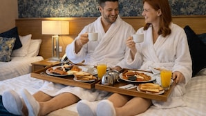 Ein Paar in einem Hotelzimmer mit Frühstückswagen, Bett, Kissen und Lampe.