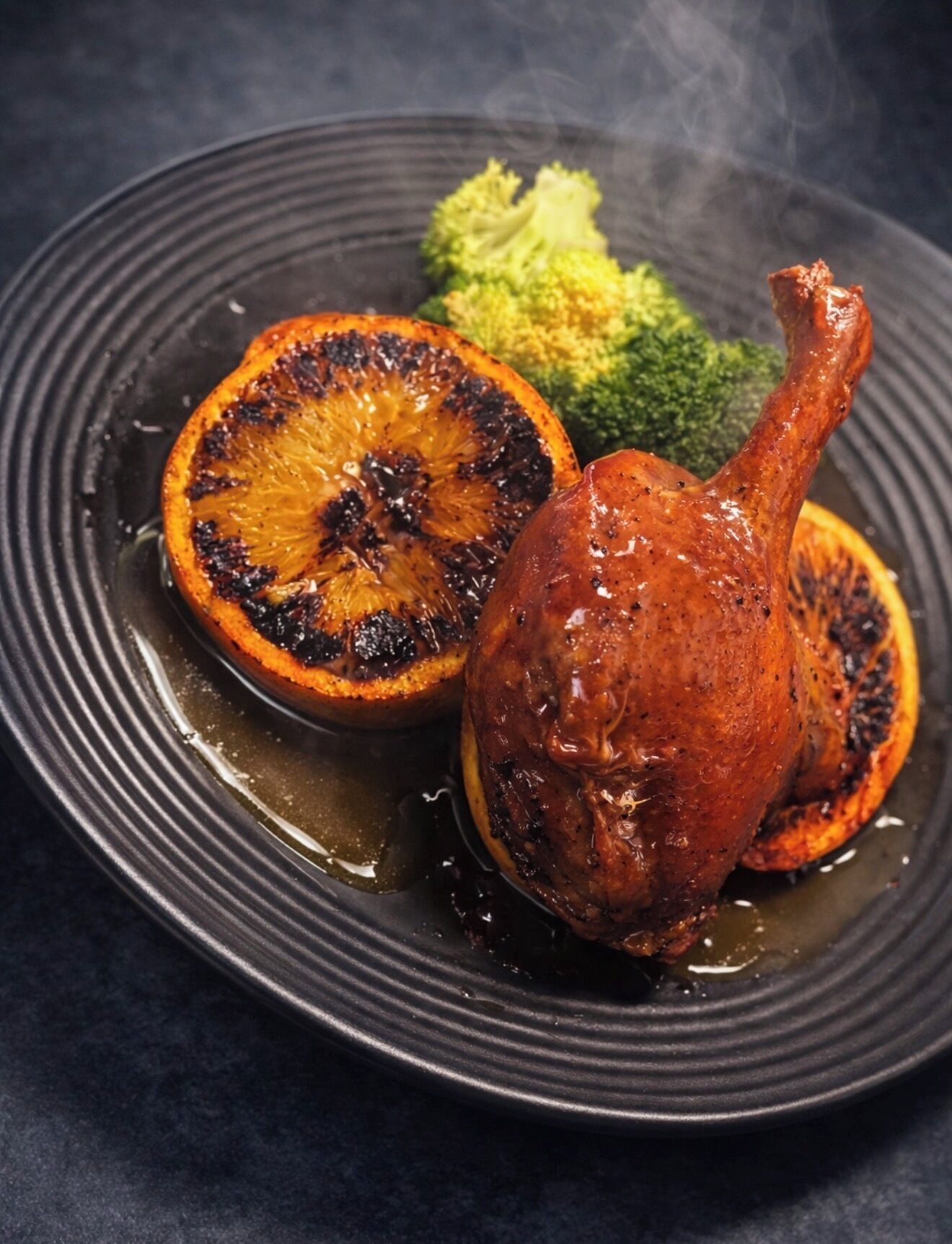 Un pato asado con piel crujiente, servido con una rodaja de naranja a la parrilla y brócoli al vapor sobre un plato oscuro.