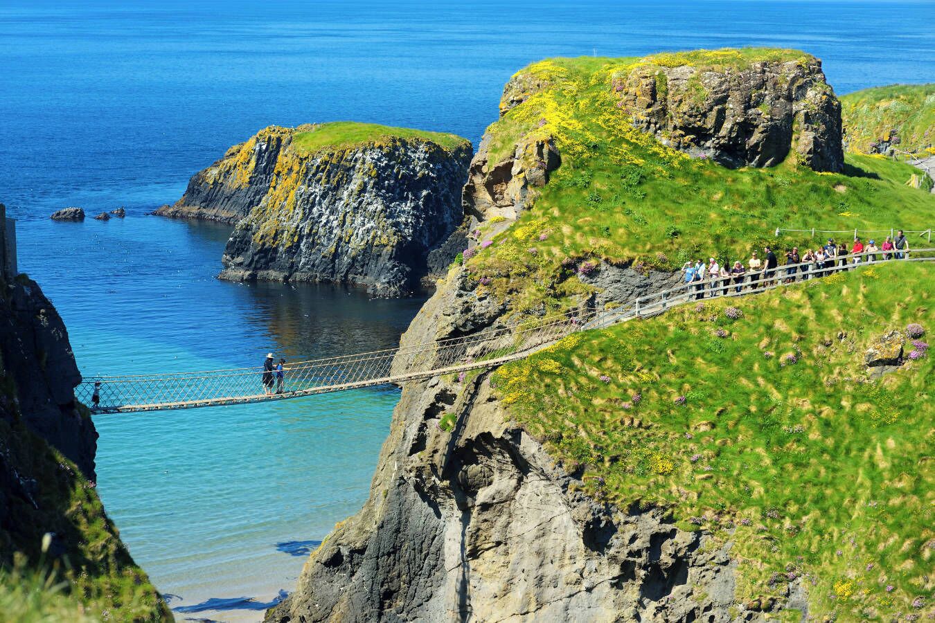 A coastal landscape with a rope bridge over a cliff, clear blue water, and a grassy area with people.