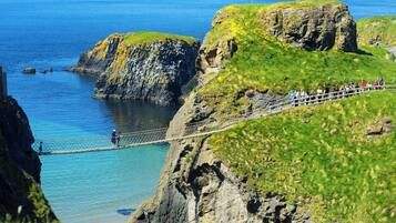 A coastal landscape with a rope bridge over a cliff, clear blue water, and a grassy area with people.