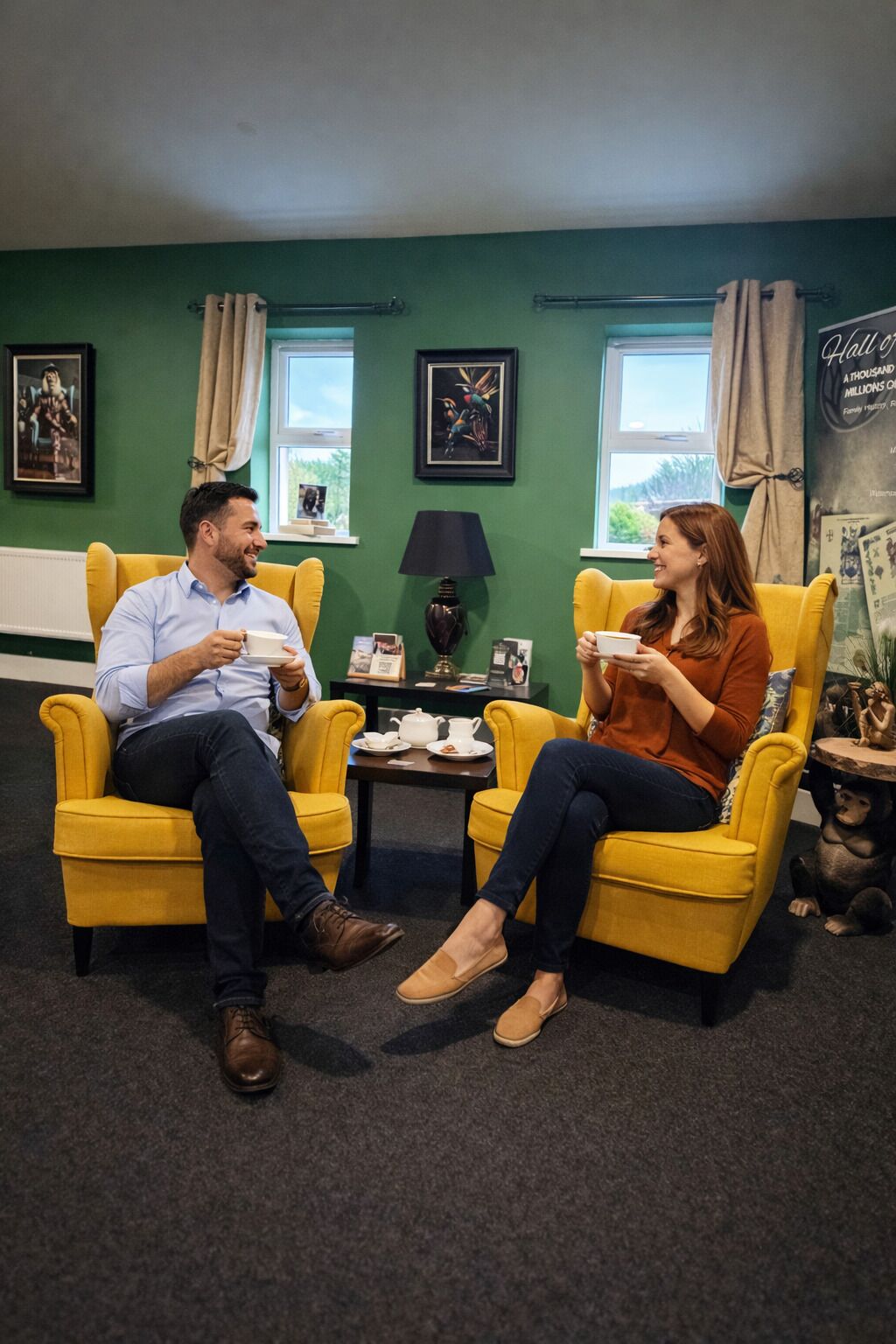 Two people seated in yellow armchairs in a room with green walls, a small table with a teapot, and framed pictures on the wall.