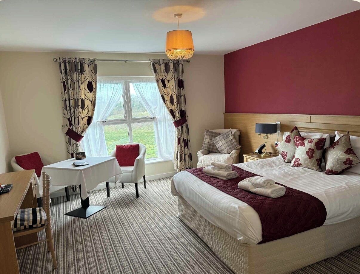 Quarto de hotel com cama, secretária, duas cadeiras, mesa, abajur e janela com cortinas.