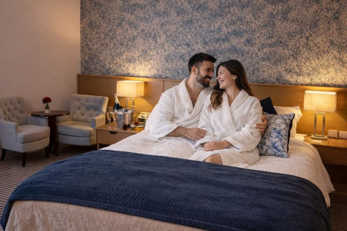 A hotel room with a large bed, two armchairs, a small table with a vase, and a patterned wall.