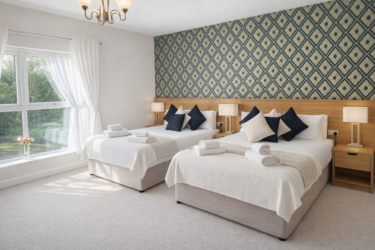 A hotel room with two beds, a large window with curtains, a patterned wall, and a chandelier.