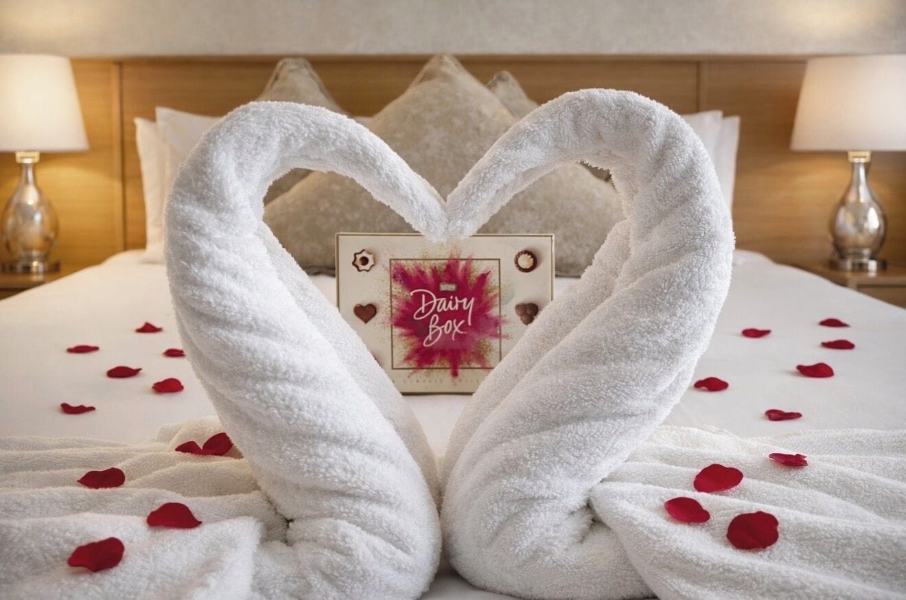Una cama con toallas blancas dispuestas en forma de corazón, un letrero 'Dairy Box' y pétalos de rosa esparcidos sobre la cama.