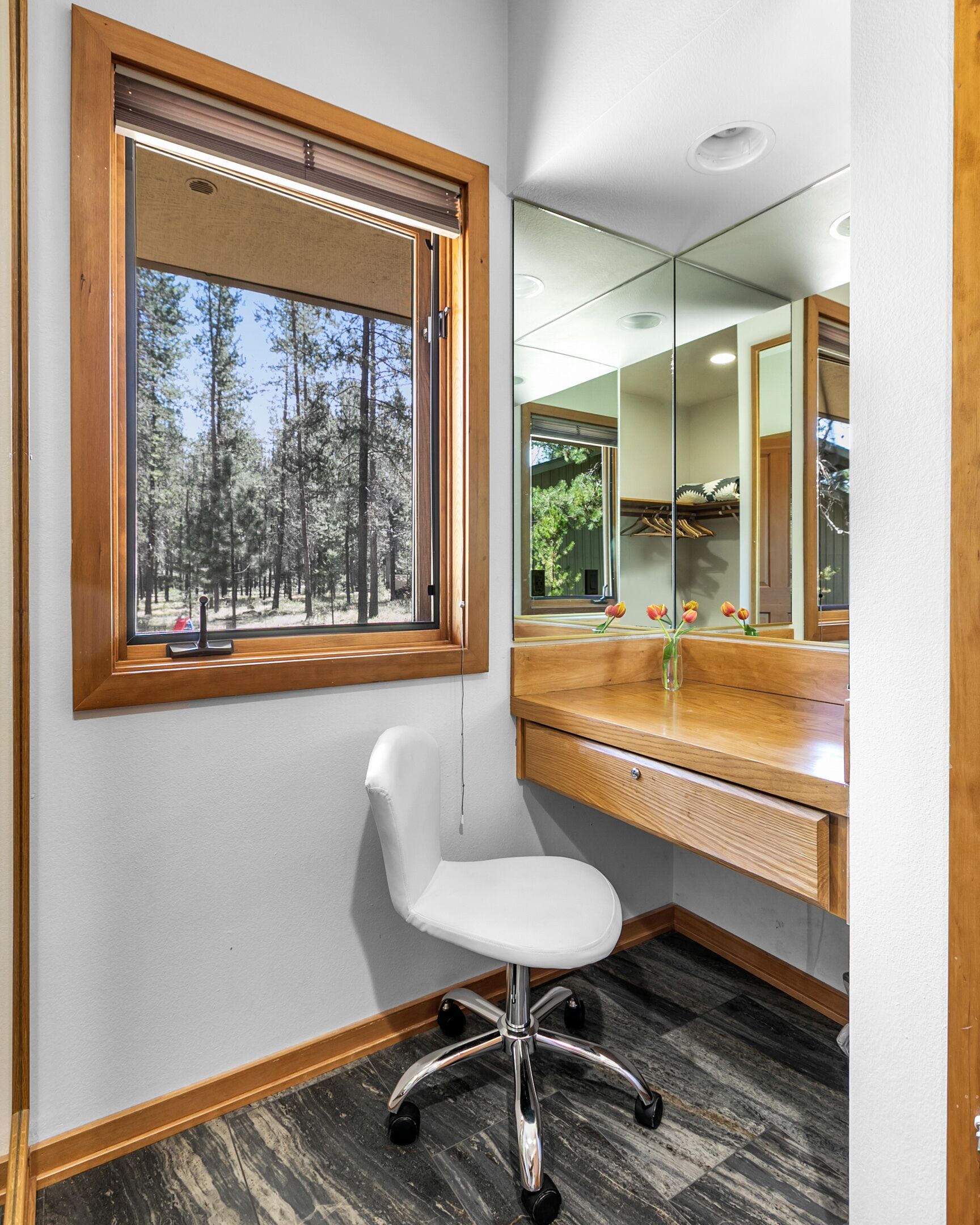 A corner room with a wooden desk, a white chair, a large mirror, and a window with a view of trees.