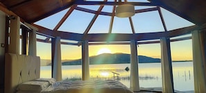 Una habitación con un tragaluz grande, una cama y vistas a un lago y montañas al atardecer.