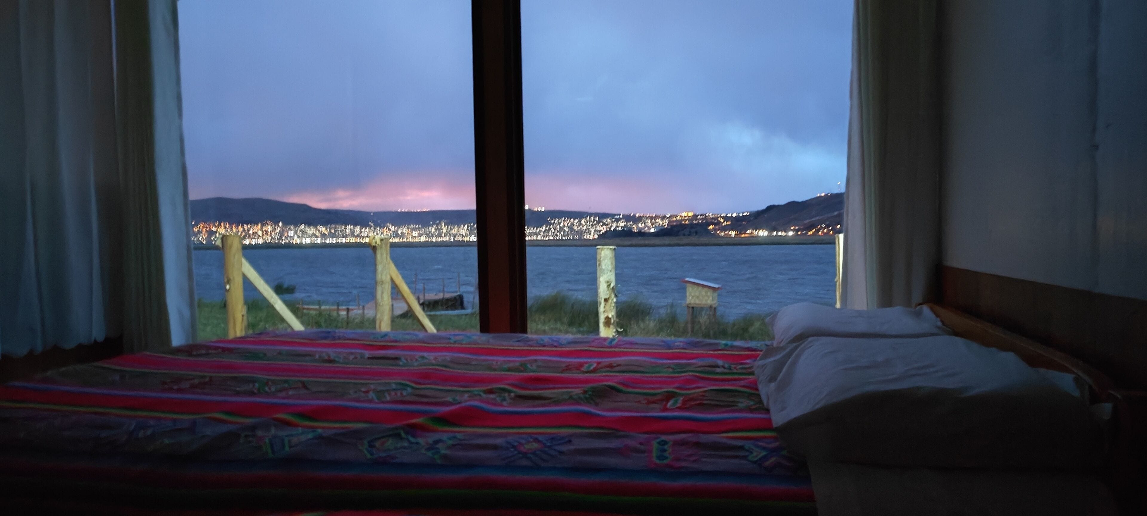 Una cama con una colcha colorida, vista de un paisaje urbano al otro lado del agua, y una valla de madera en primer plano.