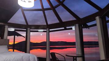 A room with a bed, a table, and chairs, with a view of a sunset over a body of water.
