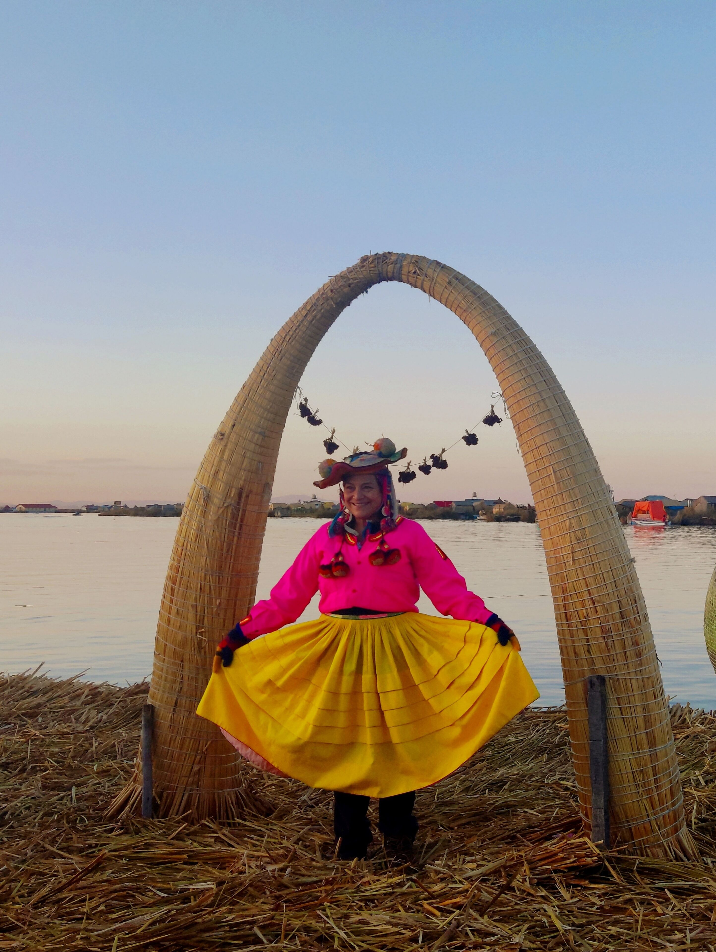 A person standing in front of a straw arch structure with a body of water and buildings in the background.
