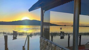 A view from a covered outdoor area with a reflection of the sunset on a calm body of water, surrounded by reeds and a distant mountain range. - Vanalux Titicaca (Puno)